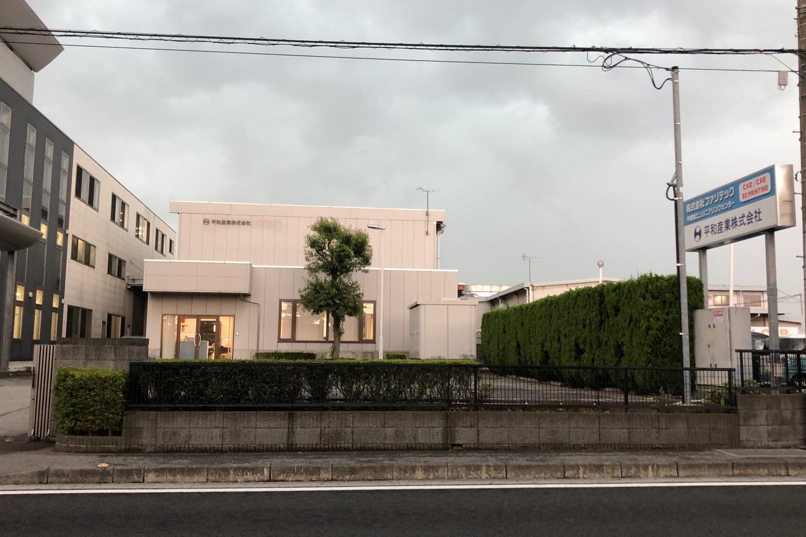 植え込みの奥にある、ベージュの外壁の工場と社名の看板